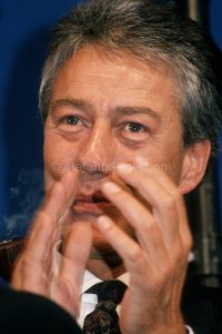 Mann mittleren Alters mit grau meliertem Haar trägt Hemd und gemusterte Krawatte, hält Zigarette und gestikuliert.