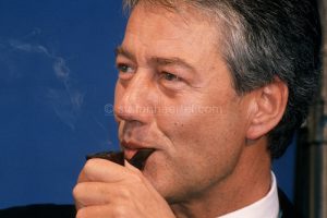 Älterer Mann mit grauem Haar raucht Pfeife vor blauem Hintergrund, trägt Sakko und weißes Hemd.