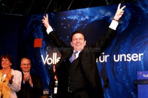 Mann in Anzug zeigt Peace-Zeichen vor blauem Hintergrund mit Text, umgeben von applaudierenden Menschen.