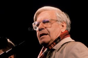 Älterer Mann mit heller Jacke und kariertem Schal spricht in Mikrofon vor dunklem Hintergrund.