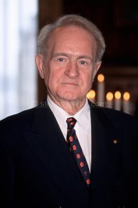 Älterer Mann mit hellem Haar in dunklem Anzug, weißem Hemd und gemusterter Krawatte vor Kerzen.